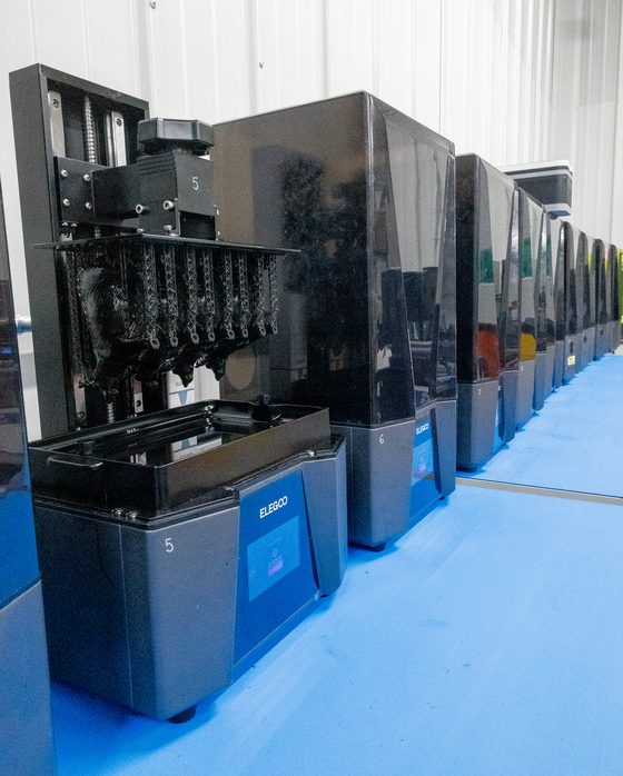 A resin printer open mid run, its build plate full of printed parts, in a row of resin machines
